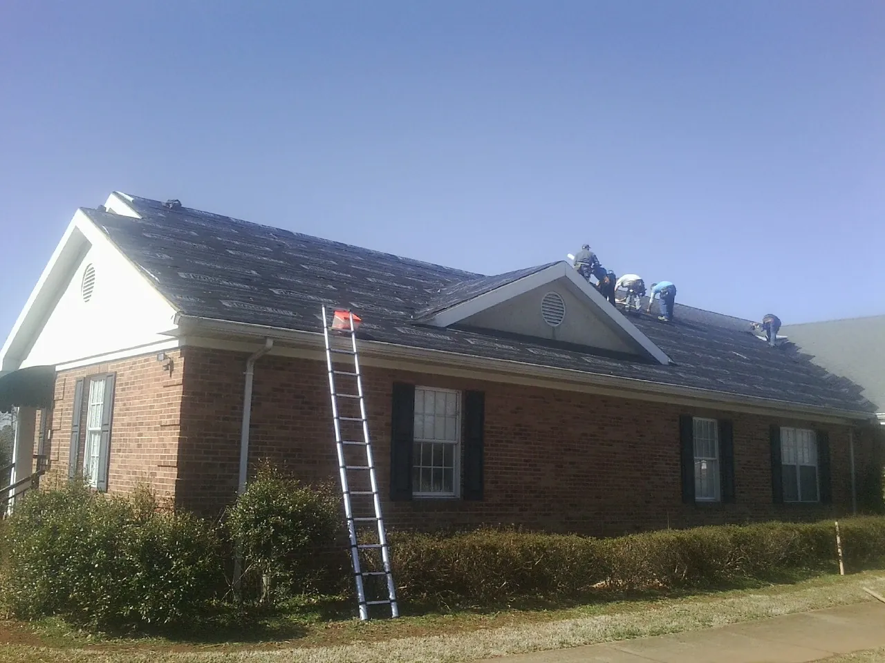 Crew tearing shingles off a brick ranch roof with a ladder set — Metal Roof Repair work by our crew in Booneville