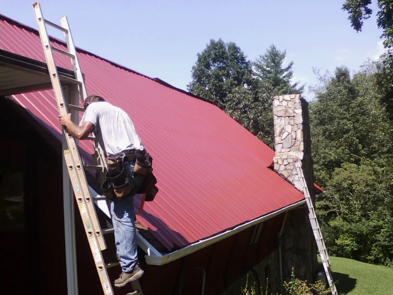 Roofer on a ladder beside a red metal roof and stone chimney — Soffit & Fascia Repair by Roofing Brothers in Booneville